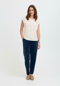 Krämfärgad blus med veckad halsringning och korta ärmar, kombinerad med marinblå kordbyxor och beige slip-on skor. En enkel vit bakgrund.