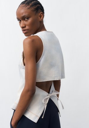 Frau mit geflochtenem Haar trägt ein ärmelloses, blass gestreiftes Top mit offenem Rücken, das mit einer Schleife gebunden ist, kombiniert mit dunkler Hose, vor weißem Hintergrund.