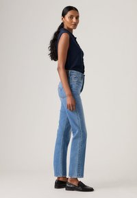 Γυναίκα με μακριά σκούρα μαλλιά στέκεται πλευρικά φορώντας αμάνικη μπλούζα ναυτικού χρώματος, ανοιχτόχρωμα μπλε τζιν και μαύρα μοκασίνια μπροστά σε απλό φόντο.