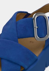 Μπλε σουέντ σανδάλι με σχεδιασμό ζωνών που διασταυρώνονται. Διαθέτει ασημένια αγκράφα και υφασμένο μπεζ εσωτερικό πέλμα, τοποθετημένο σε σταθερή μπλε βάση πλατφόρμας.