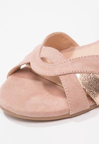 Sandale à bout ouvert avec des brides croisées en daim rose doux et un accent étroit en métal doré sur une semelle claire.