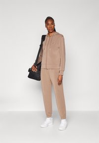 Haut à capuche beige et joggings en tissu doux, avec une fermeture éclair à l'avant et des poignets élastiques. Associé à des baskets blanches et un sac noir.