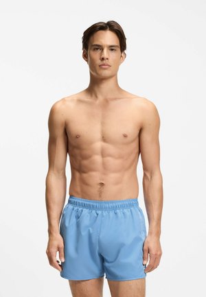 Giovane uomo con capelli corti castani che indossa pantaloncini da bagno azzurri, in piedi davanti a uno sfondo bianco semplice, volto frontale con espressione neutra.