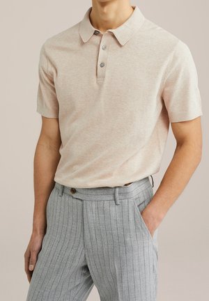 Lichtbeige poloshirt met korte mouwen en twee knopen, gecombineerd met hooggesneden grijze pantalon met krijtstreep, met één hand in de zak.