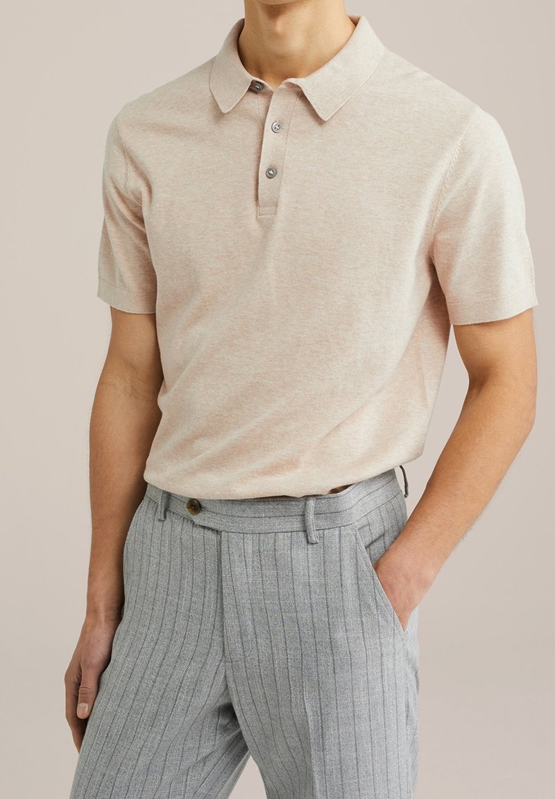 Polo manches courtes beige clair avec deux boutons, associé à un pantalon taille haute gris à fines rayures, une main dans la poche.