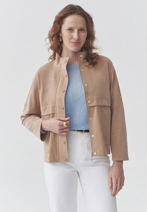 Beige jas van zacht materiaal met een hoge kraag, drukknoopsluiting en vorenzakken. Gekozen boven een blauwe trui en witte broek.