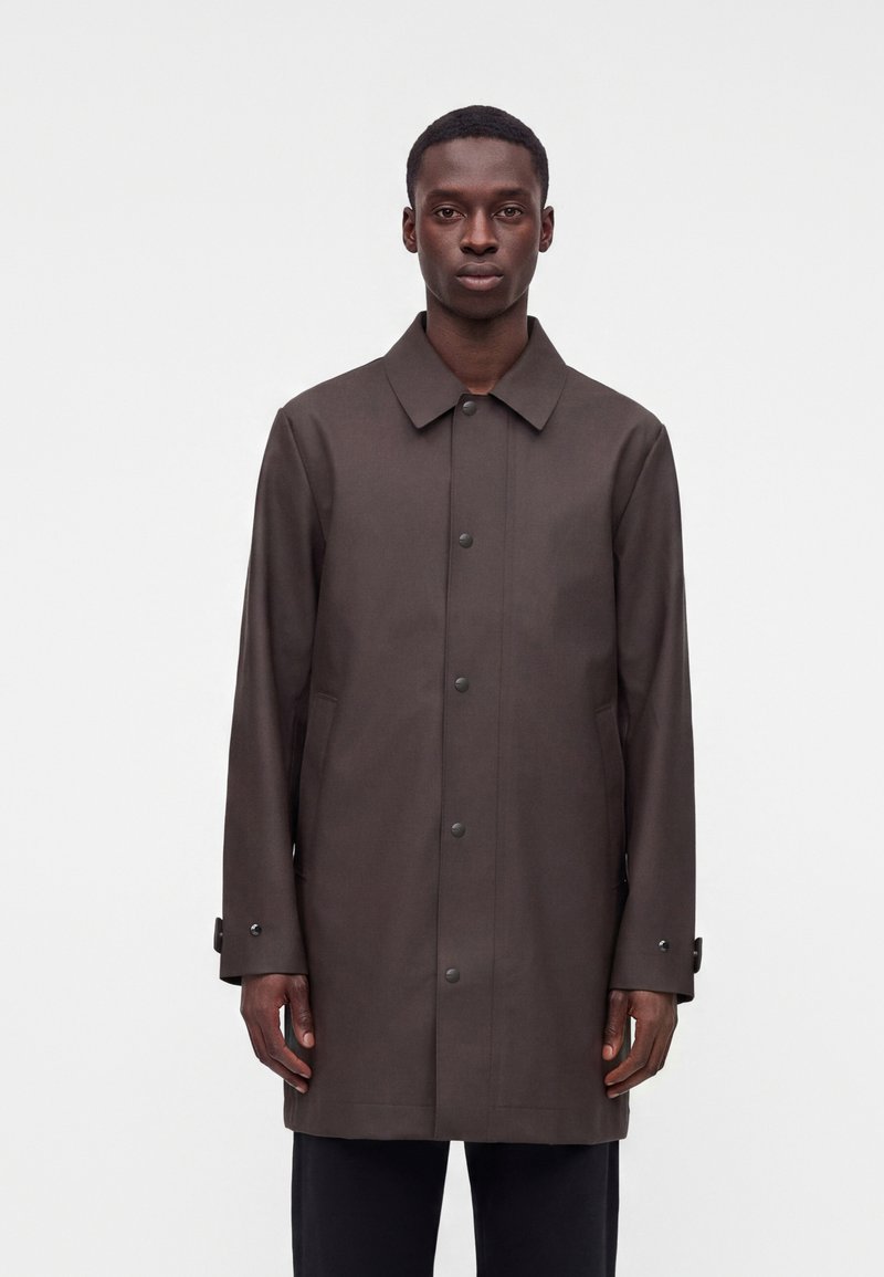 Homme debout portant un long manteau marron foncé boutonné avec un col, sur un fond clair uni.