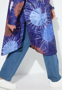 Lila oversized skjorta med flerfärgade abstrakta mönster, med tryckknappar och sidofickor, tillsammans med ljusblå jeans och vita sneakers.