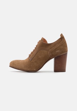 Bottine en daim marron avec lacets à l'avant, col de talon perforé et talon haut en bloc de bois épais sur fond blanc.