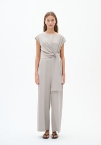 Jumpsuit grigio chiaro con maniche a cappuccio, un dettaglio a torsione davanti e un nastro in vita. Pantaloni larghi e fluidi e sandali piatti completano il look.