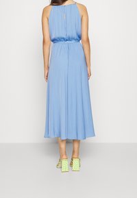 Frau in einem ärmellosen, hellblauen Midikleid mit elastischem Taillenbund und Knopf-Detail mit Ausschnitt hinten, kombiniert mit grünen karierten Blockabsatzsandalen.