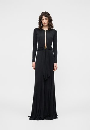 Ženska z kodrastimi lasmi, oblečena v dolgo črno obleko s sredinskim izrezom in zlato zaponko, stoji pred enostavnim svetlim ozadjem.