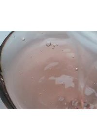 Rozenwater wordt in een heldere glazen schaal gegoten, waardoor er rimpelingen ontstaan. De vloeistof heeft een lichtroze tint met kleine luchtbellen die zichtbaar zijn.
