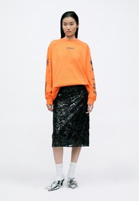 Oranger übergroßer Sweatshirt mit gedruckten Designs, kombiniert mit einem schwarzen Pailletten-Bleistiftrock. Silberne spitz zulaufende Schuhe und weiße Knöchelsocken.