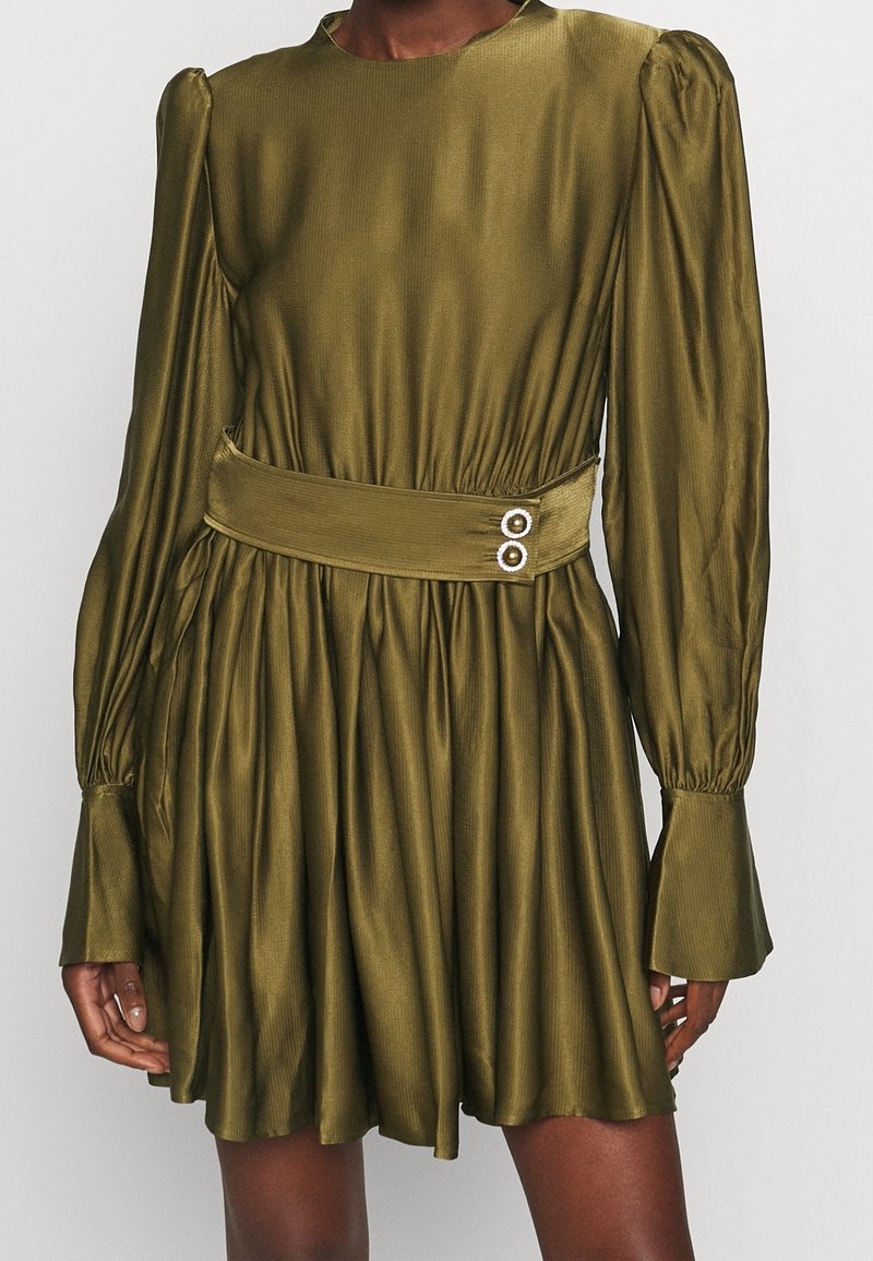 Robe vert olive avec manches longues bouffantes, large ceinture avec deux boutons décoratifs, et jupe évasée portée par une personne.
