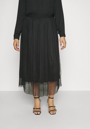 Kobieta w czarnej plisowanej spódnicy midi z przezroczystą nakładką i czarnych sandałach na wysokim obcasie z paskami, stojąca na jednolitym tle.