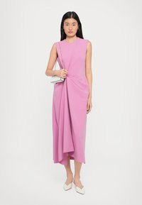 Ροζ αμάνικο φόρεμα με στριφτό σχέδιο, ελαφρύ ύφασμα και μήκος μέχρι το γόνατο. Συνδυασμένο με μια λευκή τσάντα και κομψά παπούτσια.