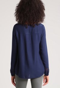 Camicia a maniche lunghe blu navy con una vestibilità comoda, caratterizzata da dettagli in pizzo sulle maniche e un orlo leggermente curvo. Tessuto morbido e leggero.