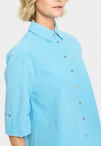 Camicia azzurro chiaro realizzata in tessuto testurizzato, con chiusura a bottoni, colletto classico e maniche arrotolate con bottoni in tonalità oro.
