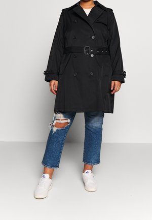 Osoba ubrana w czarny płaszcz trenck z paskiem, podarte niebieskie jeansy i białe trampki, stojąca na tle jasnego, jednolitego tła.