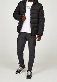 Schwarze Steppjacke mit Reißverschlusstaschen, weißes T-Shirt, dunkelgraue Cargohose und schwarze Sneakers mit weißen Akzenten.