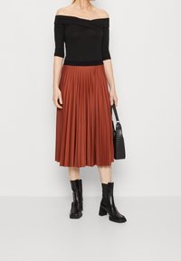 Černý top s odhalenými rameny a tříčtvrtečními rukávy v kombinaci s plisovanou midi sukní v rezavě červené barvě a černými kotníkovými botami. Součástí je i strukturovaná kabelka.