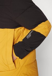 Chaqueta de plumas en negro y mostaza. Presenta una textura acolchada, puños elásticos y un detalle del logo en la manga.