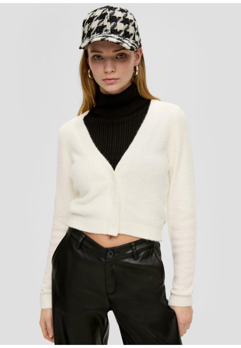 Weißer, kurzer Cardigan mit V-Ausschnitt und Knopfverschluss, über einem schwarzen, gerippten Rollkragenpullover, kombiniert mit hochgeschnittenen schwarzen Lederhosen.
