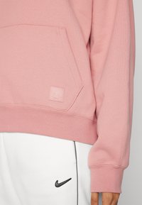 Sudadera rosa con un bolsillo frontal, con un pequeño parche de logo. La tela tiene una textura suave, combinada con pantalones de chándal blancos y un logo negro.