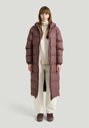 Kvinde iført en lang brun dunjakke over en hvid rullekrave sweater og vide bukser, stående mod en ensfarvet lys baggrund.