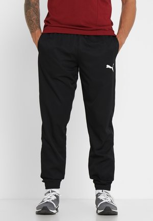 Mężczyzna w czarnych spodniach dresowych Puma i szarych butach sportowych, z rękami w kieszeniach, stoi na białym tle.