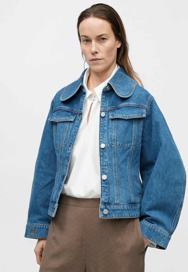 Light blue denim jacket with large collar, two chest pockets, and silver buttons. Relaxed fit with seam detailing and wide sleeves.