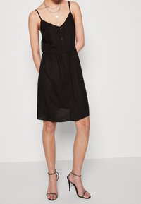 Černé šaty bez rukávů s výstřihem do V, zapínací na knoflíky vpředu a s elastickým pasem. Texturovaná látka s volným střihem, doplněná černými sandály na tenkých páscích.