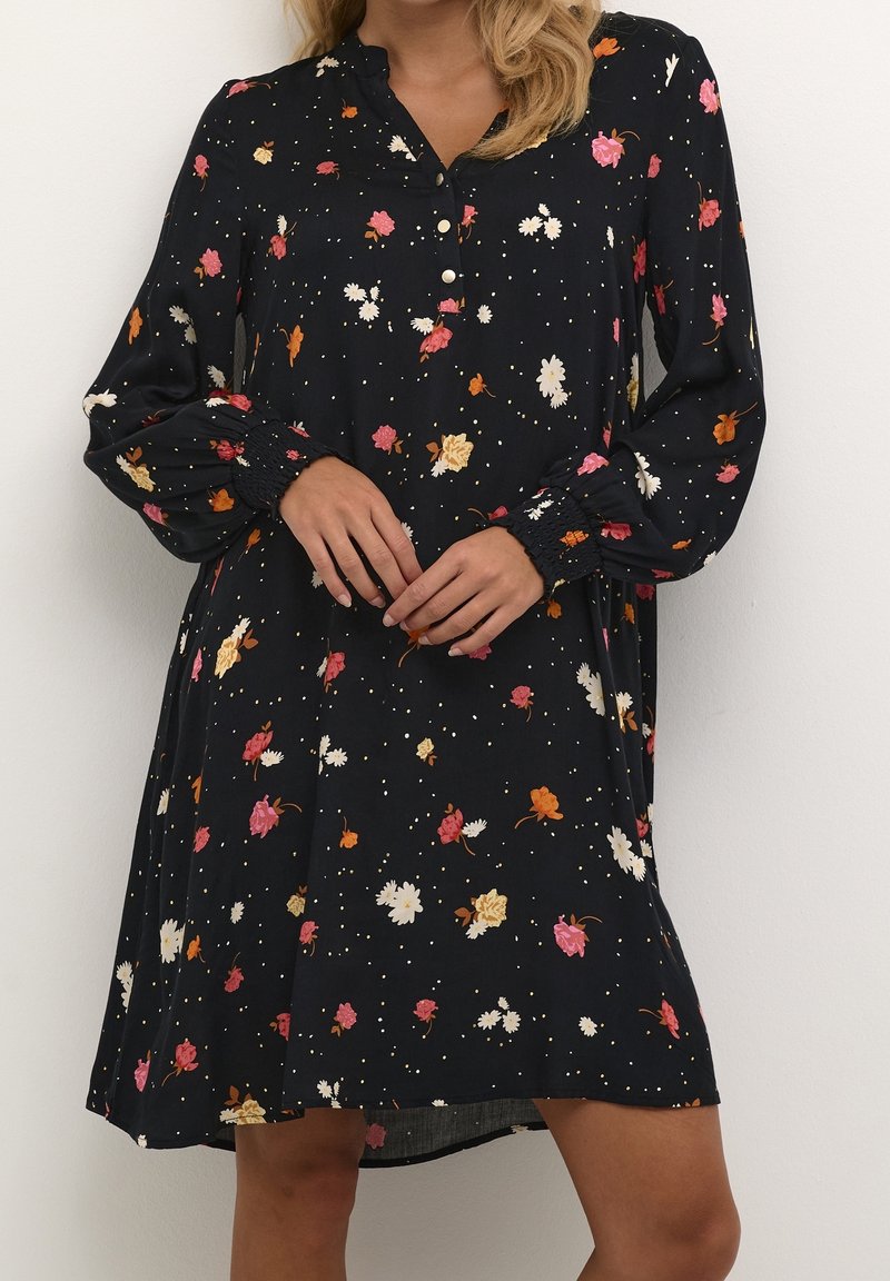 Femme portant une robe noire à manches longues avec un imprimé floral coloré et un col boutonné, mains jointes devant sur un fond blanc.