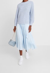 Helleblaue Strickjacke mit strukturiertem Muster, kombiniert mit einem fließenden, hellblauen Faltenrock. Weiße Sneakers und eine kleine Kettenhandtasche runden den Look ab.