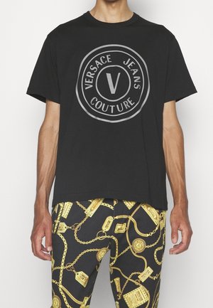 T-shirt noir à manches courtes avec le logo Versace Jeans Couture imprimé dans des cercles gris, associé à un pantalon noir avec un motif de chaîne et d'étiquette dorés.