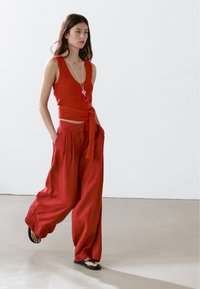 Röd stickad ärmlös topp med knyt i midjan, tillsammans med lösa, långa röda byxor. Modellen har på sig flip-flop-sandaler och står på ett ljust grått golv.