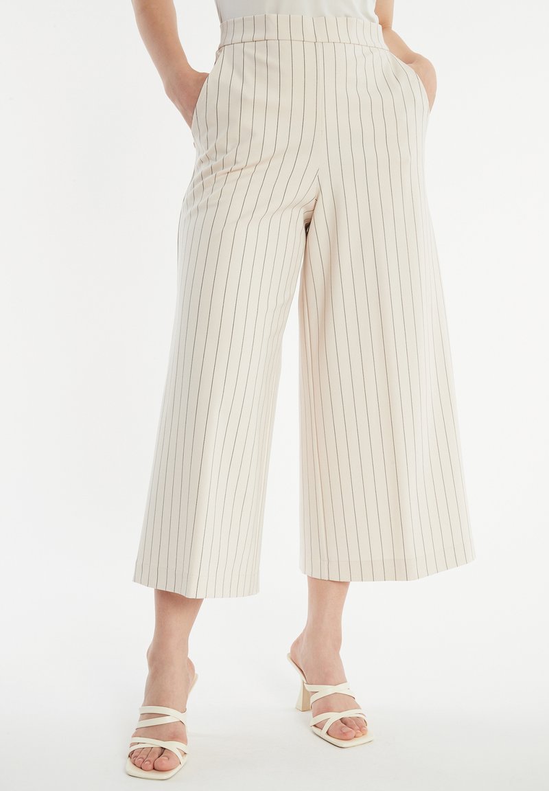 Beige weite Hose mit vertikalen Nadelstreifen, hoher Taille, zwei seitlichen Taschen und einer glatten Stofftextur. Kombiniert mit weißen Riemchenschuhen.