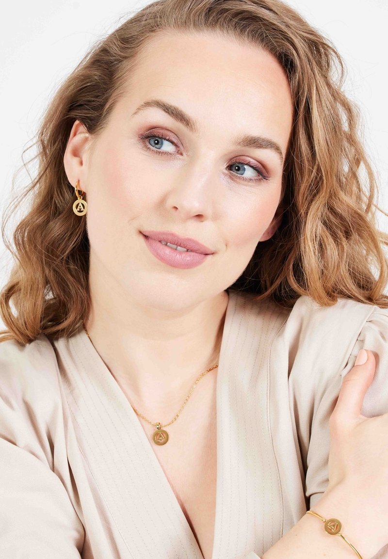 Gouden sierset met ronde oorbellen, een ketting met een hanger en een armband, allemaal met een logo-detail. Draag het met een lichtbeige top.