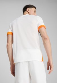 Camisa deportiva blanca con acentos naranjas en las mangas. Fabricada de tela de malla, presenta un cuello clásico y un diseño de corte amplio.