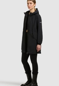 Mujer con una chaqueta larga negra con capucha, leggings negros y botines negros, de pie frente a un fondo liso.