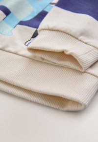 Polsino di felpa color crema con texture a coste e finitura liscia, con un motivo grafico colorato sul tessuto principale.