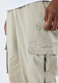 Pantalones cargo beige con un tejido texturizado, que cuentan con un bolsillo lateral y un logo bordado en el muslo. El diseño incluye un corte relajado.