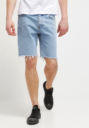 Uomo che indossa pantaloncini di jeans blu chiaro sfilacciati, t-shirt bianca e scarpe da ginnastica nere che cammina su sfondo bianco.