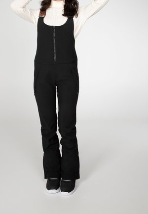 Kvinde iført sorte sneakbukser med bryststrop og lynlåslommer over en hvid, langærmet top, stående med armene løftet bag hovedet.