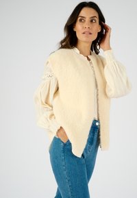 Chaleco de lana crema con cuello alto y cierre de botones, superpuesto sobre una blusa ligera y texturizada con mangas abullonadas y jeans de mezclilla.