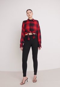 Chemise coupée à carreaux rouge et noir avec un nœud à l'avant, associée à des leggings noirs et des talons à lanières. Tissu lisse, coupe décontractée, manches longues.