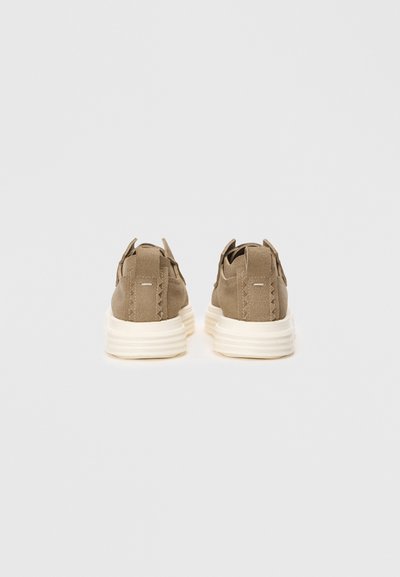 Brązowe zamszowe sneakersy o teksturowanym wykończeniu, z białą, nieco podwyższoną podeszwą i tylnymi uchwytami. Minimalistyczny design i szczegóły szwów.