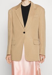Persona con un blazzer beige de un solo botón sobre una blusa negra y una falda de satén rosa brillante con detalles atados en el lateral.
