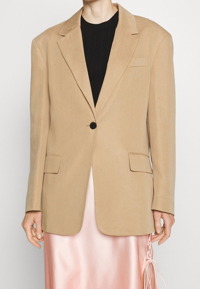 Persona con un blazzer beige de un solo botón sobre una blusa negra y una falda de satén rosa brillante con detalles atados en el lateral.